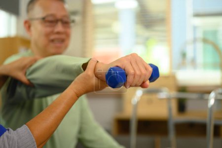 Foto de Fisioterapeuta ayudando a un hombre mayor a hacer ejercicio con mancuerna ligera en casa. - Imagen libre de derechos