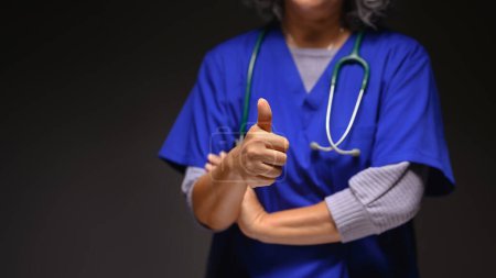 Foto de Médico maduro en uniforme médico mostrando pulgares hacia arriba sobre fondo negro. Concepto de salud. - Imagen libre de derechos