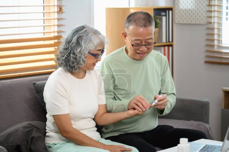 Téléchargez les photos : Homme âgé souriant aidant sa femme à vérifier le taux de sucre dans le sang à la maison. Concept de soins de santé. - en image libre de droit