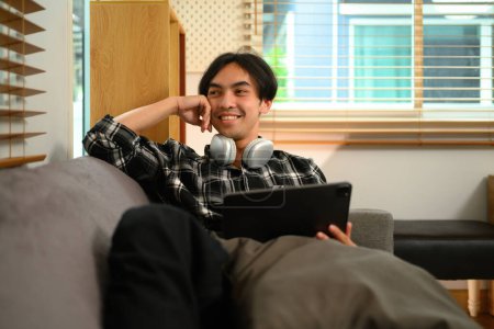 Foto de Feliz joven asiático hombre mirando hacia otro lado mientras está sentado con la tableta digital en el sofá en casa. - Imagen libre de derechos