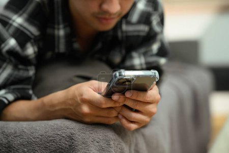 Foto de Recortado disparo de hombre joven leyendo mensajes en el teléfono móvil acostado en el sofá en casa. - Imagen libre de derechos