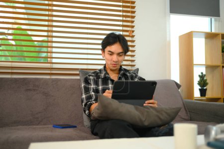 Foto de Sonriendo Joven hombre viendo vídeo en la tableta digital mientras se relaja en un sofá en casa. - Imagen libre de derechos