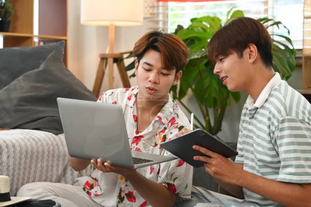 Foto de Feliz pareja gay joven planeando viaje de vacaciones, utilizando el ordenador portátil para reservar un hotel en el sitio web. - Imagen libre de derechos