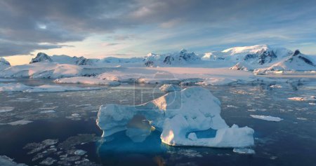 Sunset Antarctic iceberg floating in icy ocean. Snow-capped mountains in background. Fly over the untouched wilderness of Antarctica colorful landscape. Cinematic aerial drone panorama