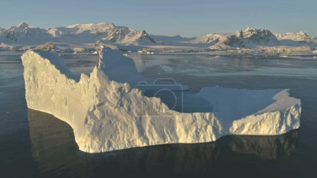 Eisberg aus nächster Nähe. Drohnenflug in der Antarktis. Verlangsamen Sie den sonnenbeschienenen Eisberg mit ovalem Wasserpool im Polarmeer. Schnee bedeckte antarktische Berge Hintergrund.