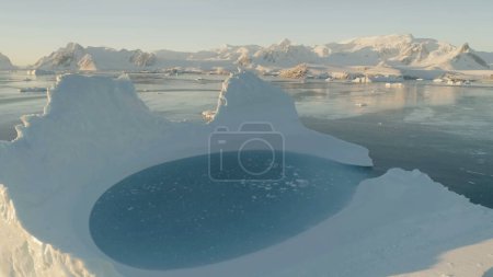 Close-up iceberg pool. Antarctica aerial view drone flight . Slow motion zoom the white iceberg with oval blue clear water pool next to the Antarctic continent. Snow covered mountains background.