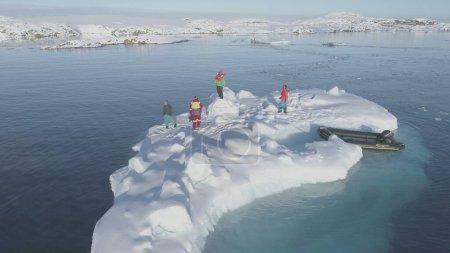 In the tranquil, icy waters of Antarctica, a team of explorers gathers on a floating ice floe, surrounded by snow-covered islands, under the bright expanse of a cloudless sky.