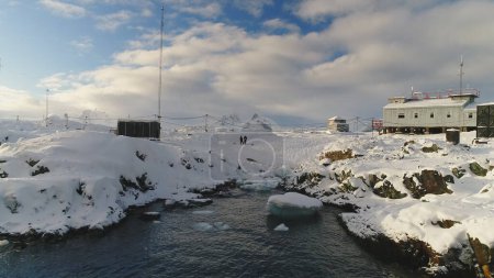 Foto de Vista de cerca, bajo vuelo de drones sobre el agua cerca de la estación polar antártica - Base Vernadsky. Gente en el paisaje de la costa oceánica. Asentamiento cubierto de nieve. Vuelo aéreo lento - Imagen libre de derechos