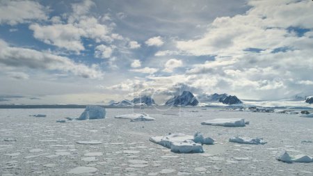 Foto de Vuelo aéreo sobre el océano Antártico. .. .. Nubes en movimiento rápido en el cielo. Drone Overview of Icebergs, Ice Pieces Floating In Sunlit Ocean (en inglés). Nieve cubierto montañas poderosas fondo. - Imagen libre de derechos