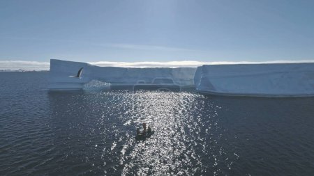 Touristenboot im antarktischen Ozean. Drohnenflug aus der Luft. Sonnenbeschienene Überblicksszene des Polarmeeres mit Eisbergen und fliegenden Vögeln. Zodiac Boot mit Menschen unter antarktischem Wasser. Exotische Reisen.
