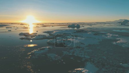 Téléchargez les photos : Coucher de soleil, océan de glace. Vol à vue aérienne par drone Antarctique. Vue d'ensemble épique et rapide du ciel couchant, au-dessus de l'océan, avec des morceaux de glace gelée, des icebergs. Voie solaire orange à la surface de l'eau. Pergélisol. - en image libre de droit