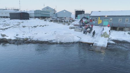 Die Menschen arbeiten am Pier der antarktischen Polarstation Wernadski. Blick auf Roboterarm zieht Boot aus. Drohnenflug vor dem Militärstützpunkt. Siedlungslandschaft am Südpol. Langsamer Flug aus der Luft.