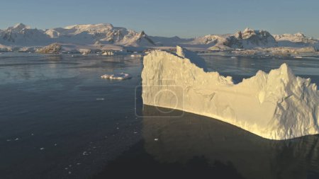Eisberg aus nächster Nähe. Drohnenflug in der Antarktis. Verlangsamen Sie den sonnenbeschienenen Eisberg mit ovalem Wasserpool im Polarmeer. Schnee bedeckte antarktische Berge Hintergrund.