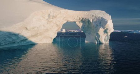 Melted ice cave arch hole iceberg towering arctic ocean. Big glacier floating cold water in sunset. Ecology, melting ice, climate change and global warming concept. Drone flight close up