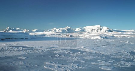 Fly over snowy mountain landscape in Antarctica. Polar frozen ocean landscape covered by snow under blue sunny sky. Discover the beauty of South Pole. Antarctica travel and exploration background