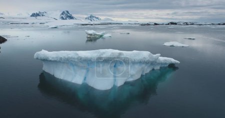 A massive iceberg floats in dark ocean water with towering mountains in the background. Antarctica icy landscapes. Polar climate change at winter day. Ecology, melting ice, global warming. Aerial shot