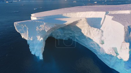 Massive iceberg drifts sunset polar ocean. Giant crashed glacier arch cave in Antarctica. Huge ice wall in pink evening light. Arctic frozen ocean landscape. Travel. Aerial top view drone flight