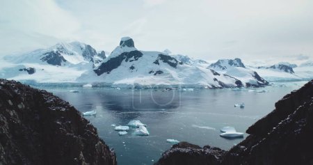 Schneebedeckte Berge atemberaubende arktische Landschaft. Schmelzende Eisberge von abgestürzten Gletschern treiben im eisigen Polarmeer. Globale Erwärmung, Klimawandel. Wilde Naturschönheiten. Kamera ragt über dunkle Felsen