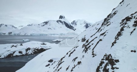Vuela sobre la roca cubierta de nieve de montaña en la Antártida. Pendiente ártica colina sobre el frío océano polar, pico de la cordillera en el fondo. Condiciones extremas de baja temperatura. Polo Sur explorar y viajar