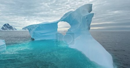 Giant melting iceberg drifts in blue ocean water. Huge high ice glacier at polar nature environment. Cold South Pole winter landscape. Wild nature. Cinematic ecology scene. Drone aerial shot