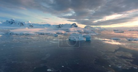 Téléchargez les photos : Hiver époustouflant Paysage de coucher de soleil aérien arctique. Beauté gelée Antarctique nature. Ciel nuageux coloré reflété dans l'eau glacée polaire de l'océan. Montagne enneigée, glacier, iceberg. Voyager, explorer - en image libre de droit