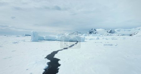 Téléchargez les photos : Survoler la colline de neige dans le paysage hivernal de l'Antarctique. Océan polaire gelé avec rivière fissurée fondue. Glacier et iceberg coincés dans la baie côtière de glace. Chaîne de montagnes en arrière-plan. Voyage et exploration dans l'Arctique - en image libre de droit