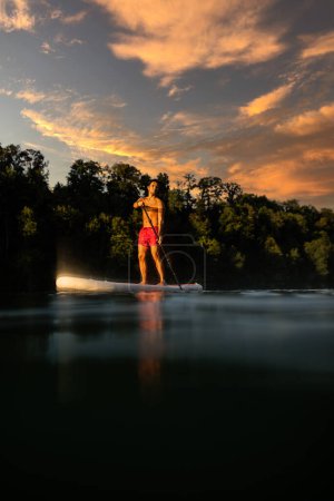 Téléchargez les photos : Beau jeune homme sur une planche à pagaie. Faire un grand exercice sur une belle rivière dans la lumière du soleil du soir chaud - en image libre de droit