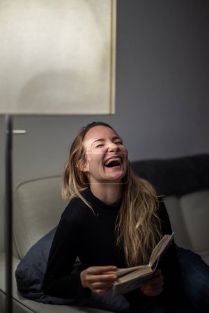 Photo for Avid reader laughing. Young attractive girl happy positive smile sit sofa home relax read book story novelty laugh humor joke - Royalty Free Image