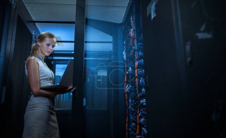 Photo for IT engineer standing amid working server racks doing routine maintenance check and diagnostics using laptop computer (color toned image) - Royalty Free Image