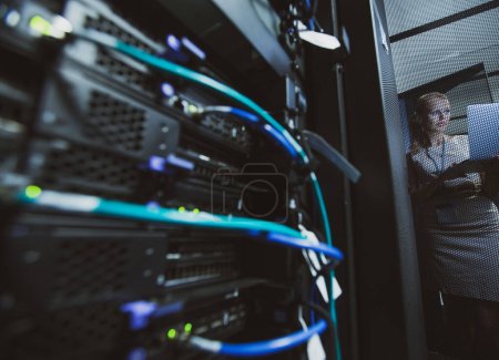 Photo for IT engineer standing amid working server racks doing routine maintenance check and diagnostics using laptop computer (color toned image) - Royalty Free Image