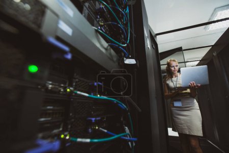 Photo for IT engineer standing before working server rack doing routine maintenance check and diagnostics using laptop - Royalty Free Image