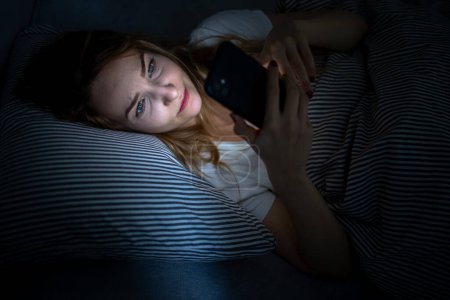Téléchargez les photos : Jeune femme au lit tenant un téléphone, fatiguée et épuisée, lumière bleue tendant les yeux, brouillant sa comptine circadienne - en image libre de droit