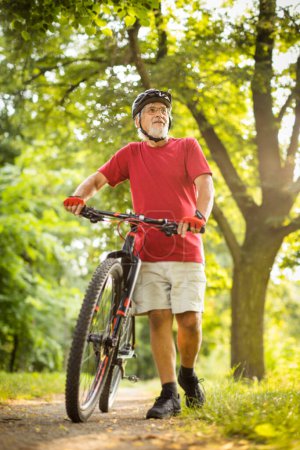 Foto de Hombre mayor con su bicicleta de montaña que va a dar un paseo más allá de los límites de la ciudad en un bosque encantador, recibiendo la dosis diaria de cardio, teniendo un descanso en un banco - Imagen libre de derechos