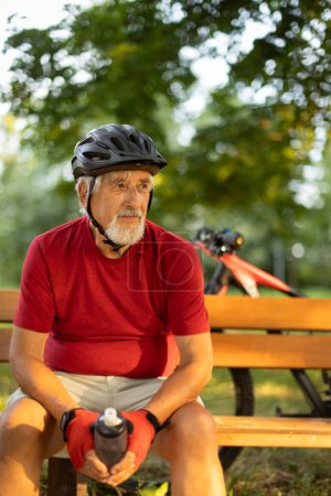Foto de Hombre mayor con su bicicleta de montaña que va a dar un paseo más allá de los límites de la ciudad en un bosque encantador, recibiendo la dosis diaria de cardio, teniendo un descanso en un banco - Imagen libre de derechos