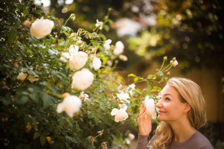 Foto de Retrato de estilo de vida de una mujer joven y bonita oliendo algunas rosas preciosas en un jardín - Imagen libre de derechos