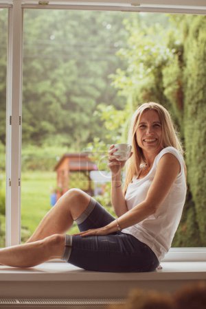 Foto de Mujer de mediana edad tomando café en casa, relajándose en una ventana con vista al jardín - Imagen libre de derechos