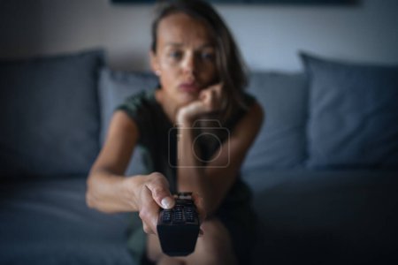 Téléchargez les photos : Jolie, jeune femme assise sur le canapé à l'intérieur à la maison et regardant la télévision - en image libre de droit