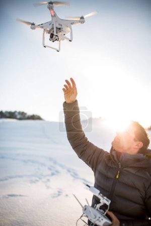 Téléchargez les photos : Jeune homme contrôlant son drone en plein air enneigé. Opérateur de drone tenant un émetteur et atterrissant avec un drone
. - en image libre de droit