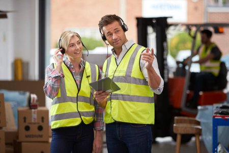 Male And Female Workers Wearing Headsets In Logistics Distribution Warehouse Using Digital Tablet