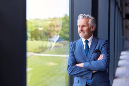 Retrato del presidente ejecutivo de Smiling Senior Businessman parado junto a la ventana dentro del edificio de oficinas moderno