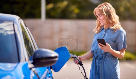 Woman Charging Electric Car With Cable Using App On Phone To Monitor Battery Level