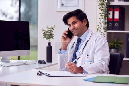 Smiling Male Doctor Or GP Wearing White Coat Sitting At Desk In Office Talking On Mobile Phone