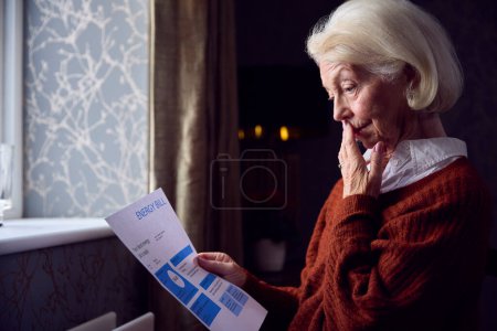 Femme âgée avec le projet de loi d'énergie des Etats-Unis essayant de garder au chaud par radiateur dans la crise d'énergie de coût de la vie