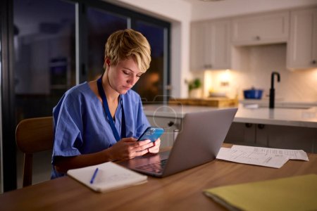 Tired Woman In Medical Scrubs Looking At Mobile Phone Working Or Studying On Laptop At Home At Night