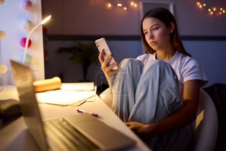 Preocupado adolescente sentado en el escritorio en el dormitorio en casa mirando el teléfono móvil por la noche