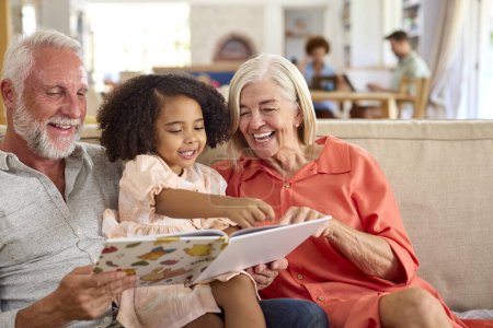 Enkelin liest mit Großeltern auf Sofa zu Hause mit Eltern im Hintergrund