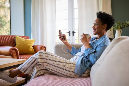 Mature Woman Relaxing On Sofa At Home With Hot Drink Looking At Mobile Phone