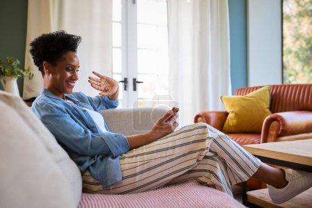 Mature Woman Relaxing On Sofa At Home Making Video Call On Mobile Phone