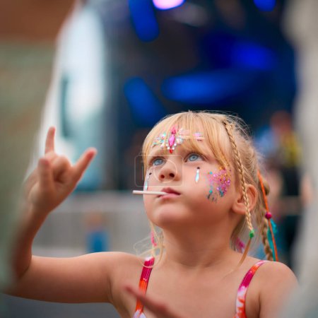 Girl Sitting On Parents Shoulders Making Rock And Roll Hand Gesture At Outdoor Summer Music Festival