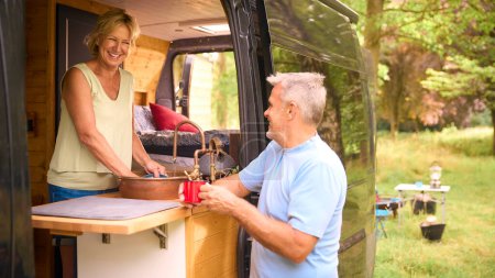 Senior Couple Enjoying Camping In Countryside Relaxing Inside RV And Doing Chores Together